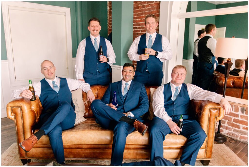 groomsmen on a leather camel colored couch in suits with drinks in their hand