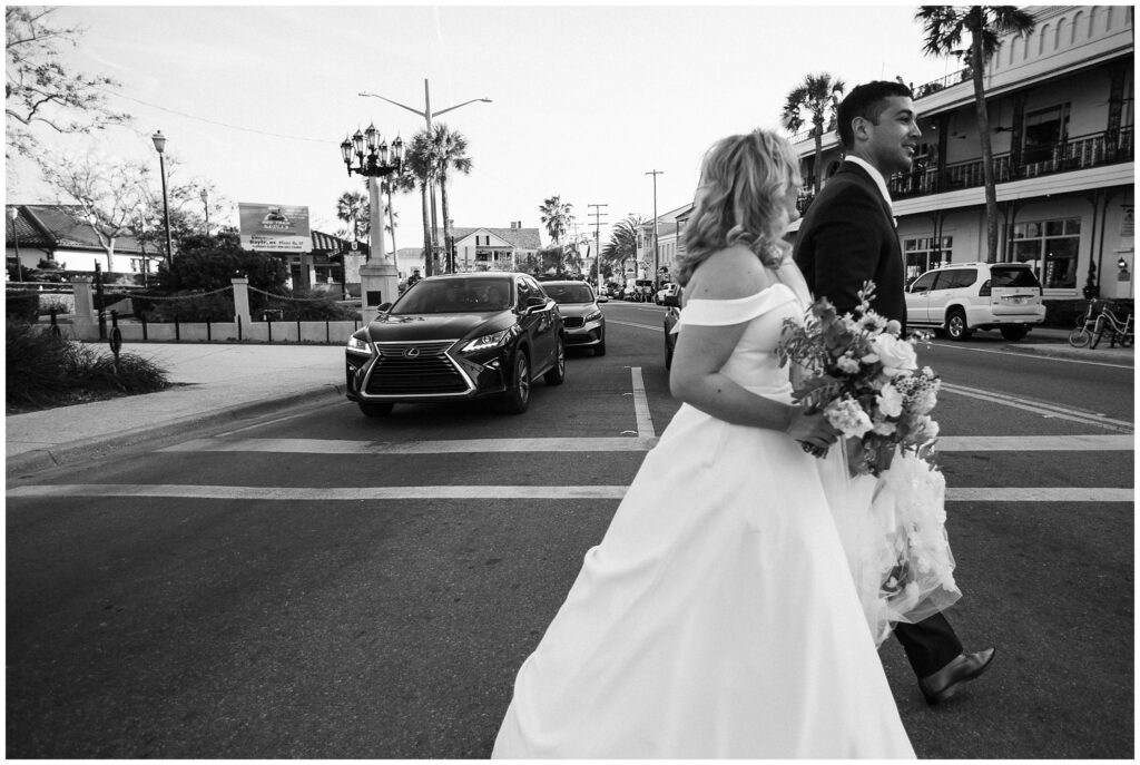 black and white photo of a bride and groom walking across a street with cars behind