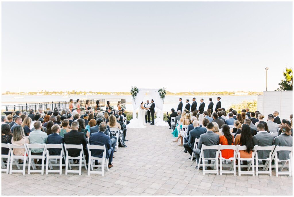 waterfront wedding ceremony set up with white chairs on a stone patio with guests in chairs and bride and groom at altar looking at each other
