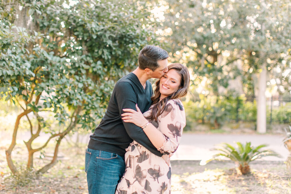 couple in front of a greenery background with man kissing the woman on the side of her forehead. 