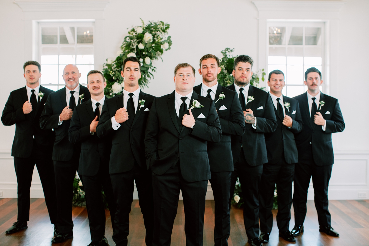 groom with eight groomsmen in black suits in front of floral arch