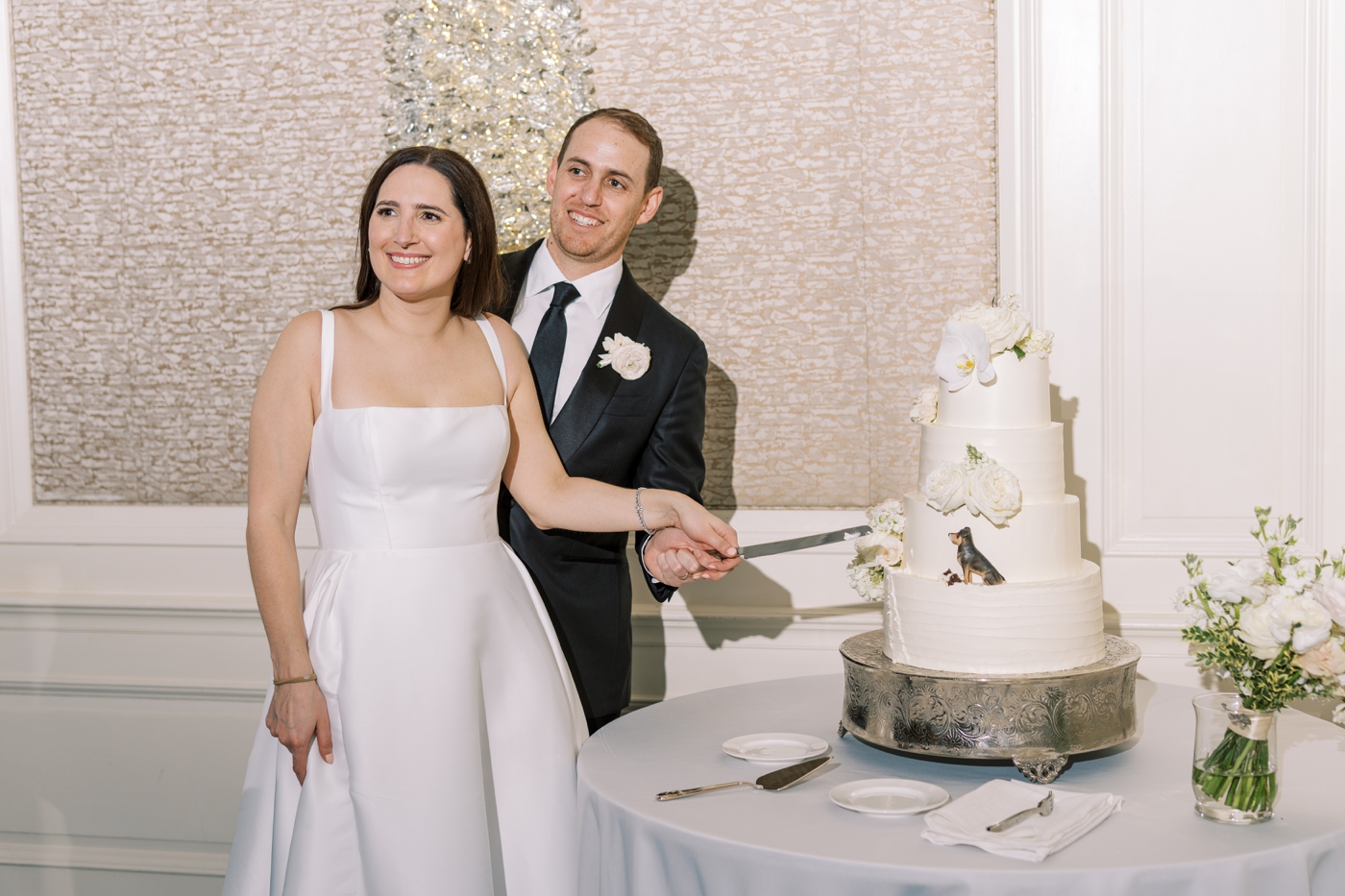 Bride and groom cutting a four-tiered white cake with flowers and a miniature figurine of their dog