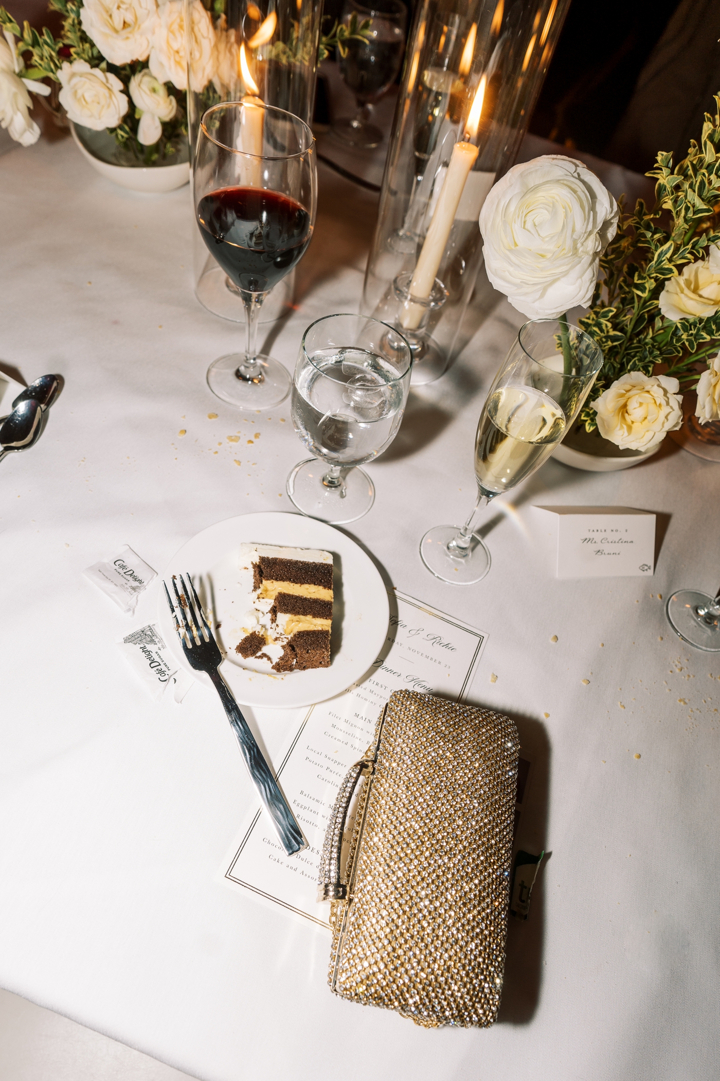 Late night wedding details including a half eaten slice of cake, glass of champagne, sugar packets, and sparkly gold clutch 